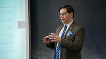 A man in a suit and tie standing in front of a blackboard giving a lecture