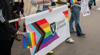 Students march with the UVM Pride banner, displaying the progress pride flag with the UVM logo in the center.