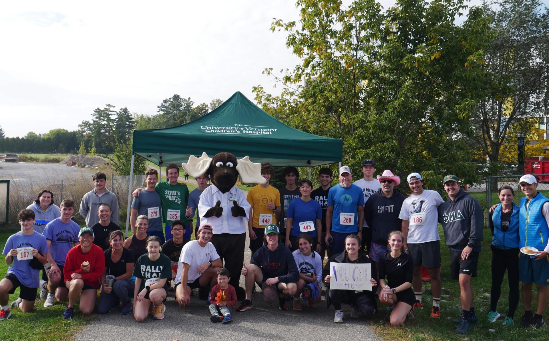 A group of 30 people, some standing and some kneeling, post for the camera. All are dressed in athletic gear and some have a white competitor number on the front of their shirts. Monty the Moose, UVM Children's Hospital mascot, poses with thumbs up with the group.