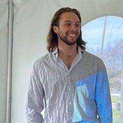 Ethan Pezzini. A person with long dark hair and a beard wearing a white dress shirt and dark pants smiles and looks away from the camera.
