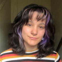 Gabby white. A person with dark hair with purple highlights and a multi-colored striped shirt looks at the camera.