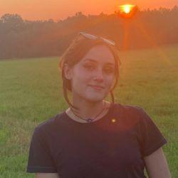 Emma Hendra. A person in a dark shirt with dark hair stands with the sun at their back.