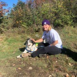 Lilah Krugman. A person with blonde hair, a purple beanie, white shirt, and black pants sits on the ground petting a white dog, both looking and smiling at the camera.