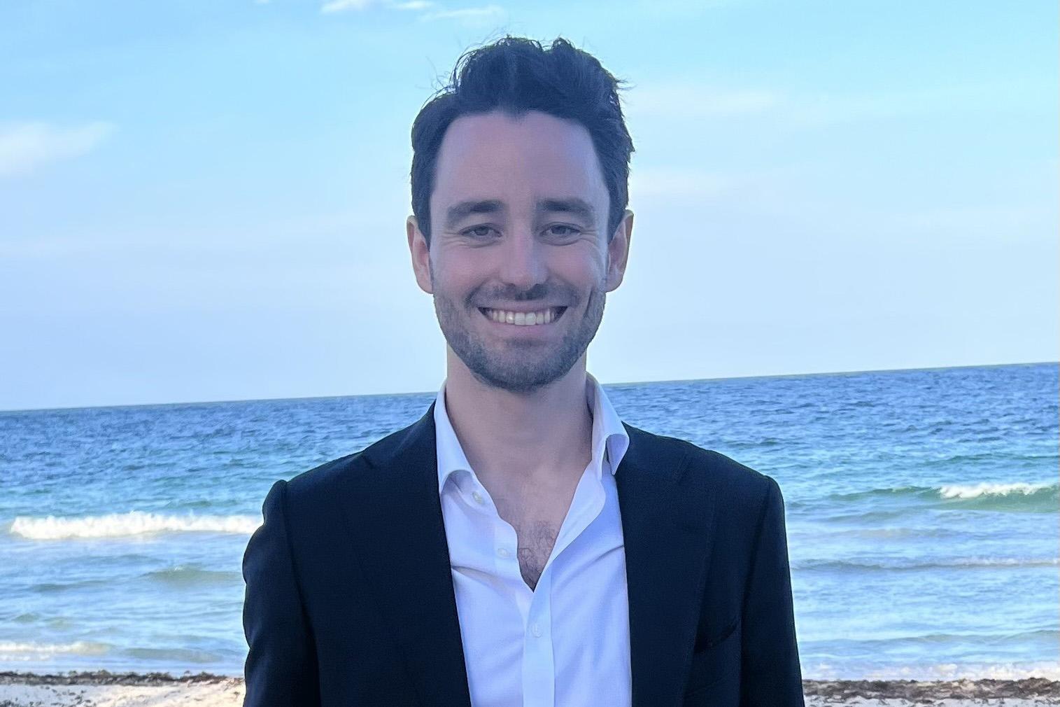 young man wearing blazer and white shirt standing on sandy beach with ocean in background