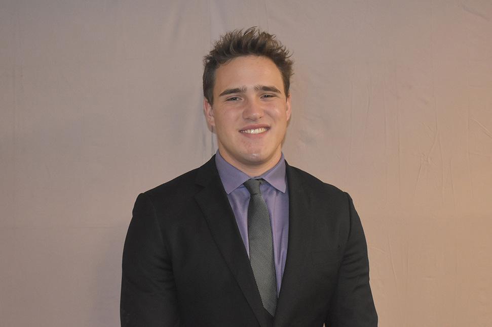 young man with short, spiky light brown hair, wearing dark gray suit and lavender shirt with tie, standing in front of wall