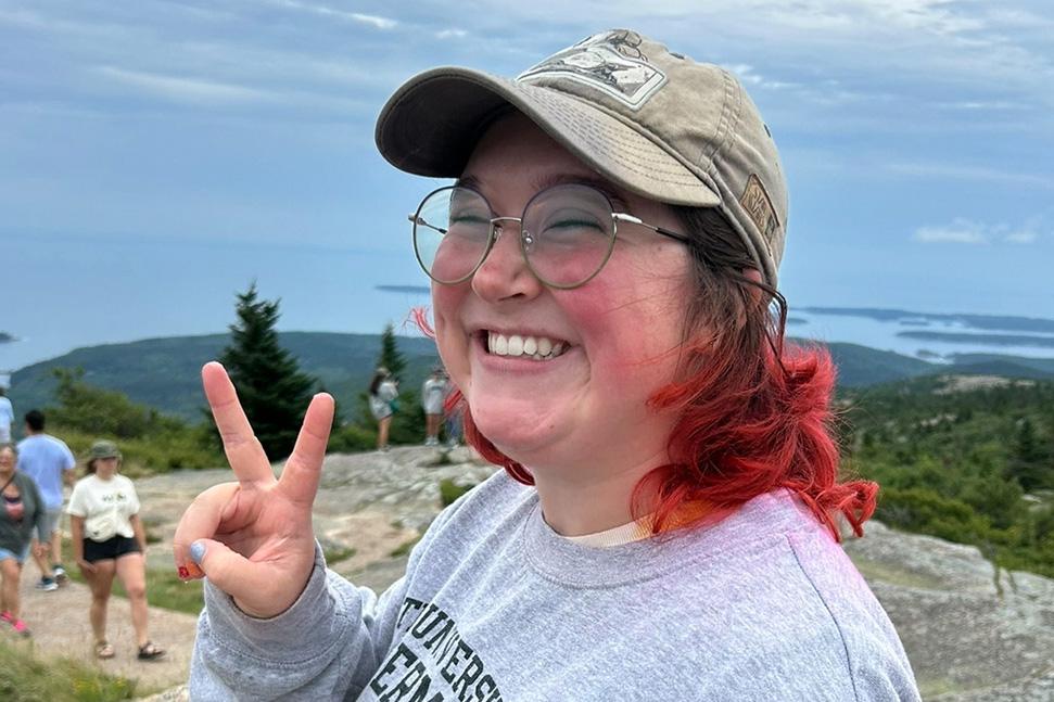 young person, smiling, with round glasses, light green ball cap, and bright red hair giving peace sign on mountaintop