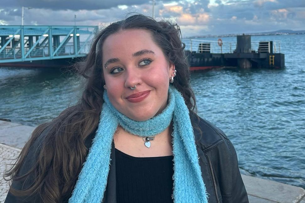 young woman with long brown hair, brown jacket, and blue scarf standing on pier by large body of water