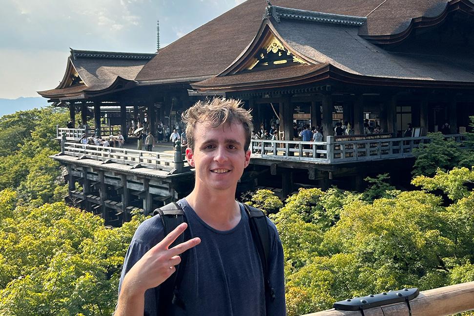 young man in blue t-shirt with light brown hair giving peace sign while standing in front of traditional Japanese building surrounded by trees