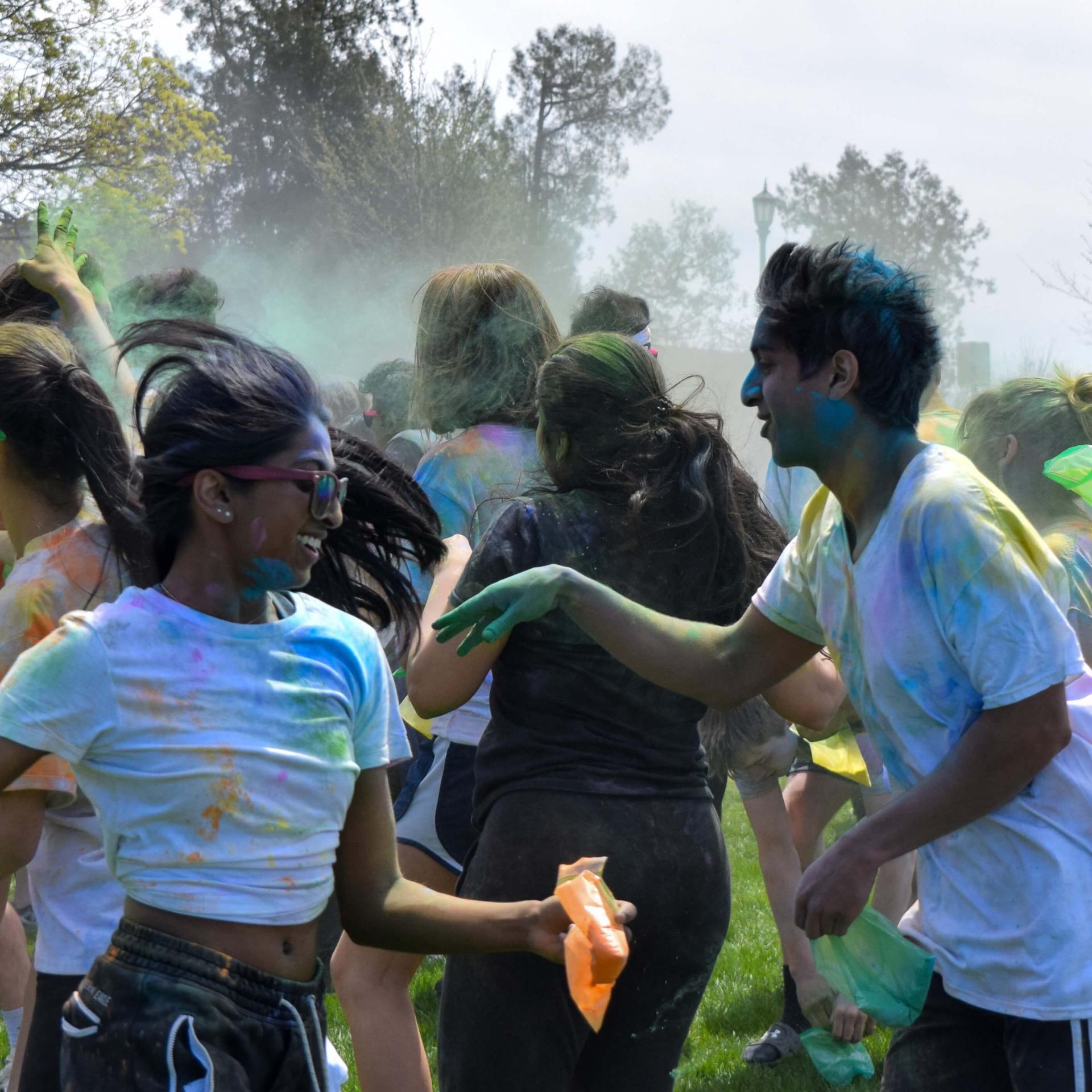 Students in white t-shirts covered in colorful paint smiling and running as they celebrate Holi