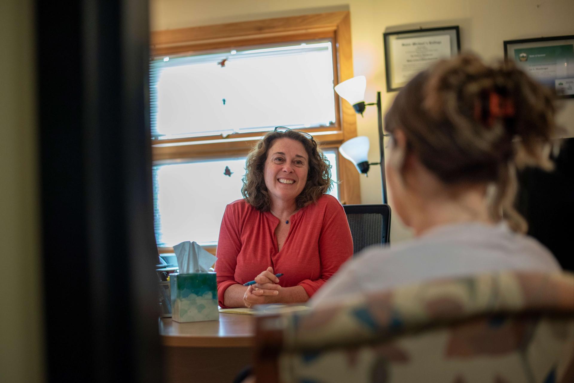 a woman sits behind a desk smiling at a student
