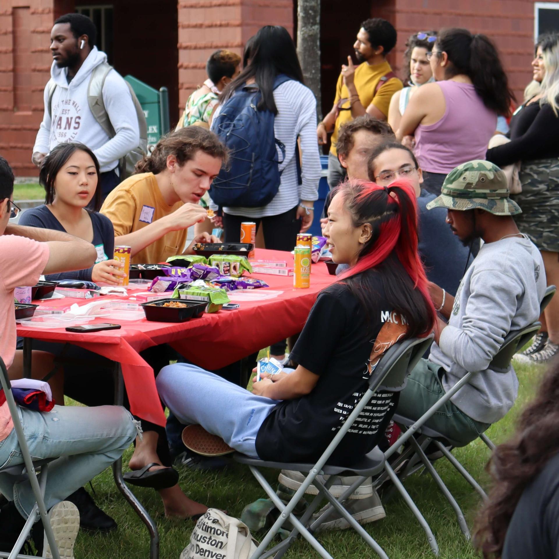 Students of different racial and ethnic backgrounds sitting at an outdoor table eating and talking while more students gather behind them.