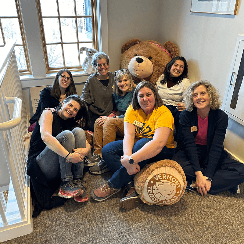 Seven people smiling and sitting on the floor of the Hope Lodge with a large teddy bear