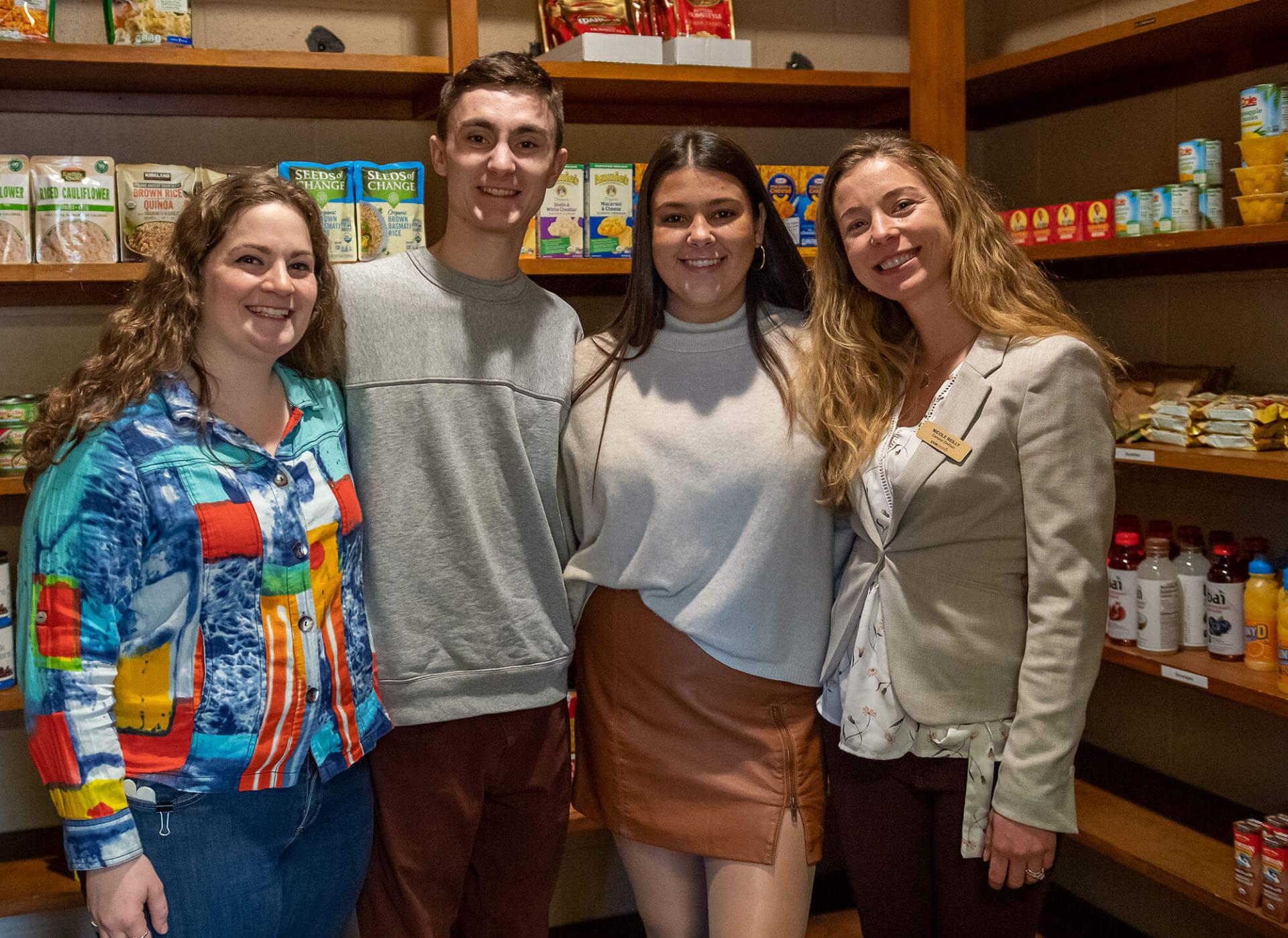 UVM staff and students stand in a group of four in front of food shelf shelves that are fully stocked.