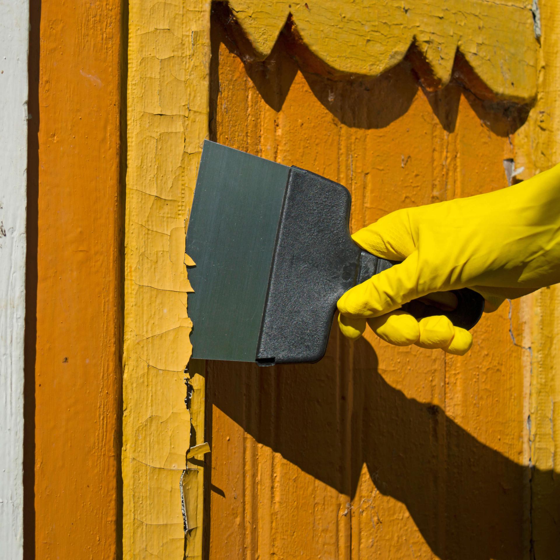 A rubber gloved hand with a scraper removes yellow paint from the side of a building.