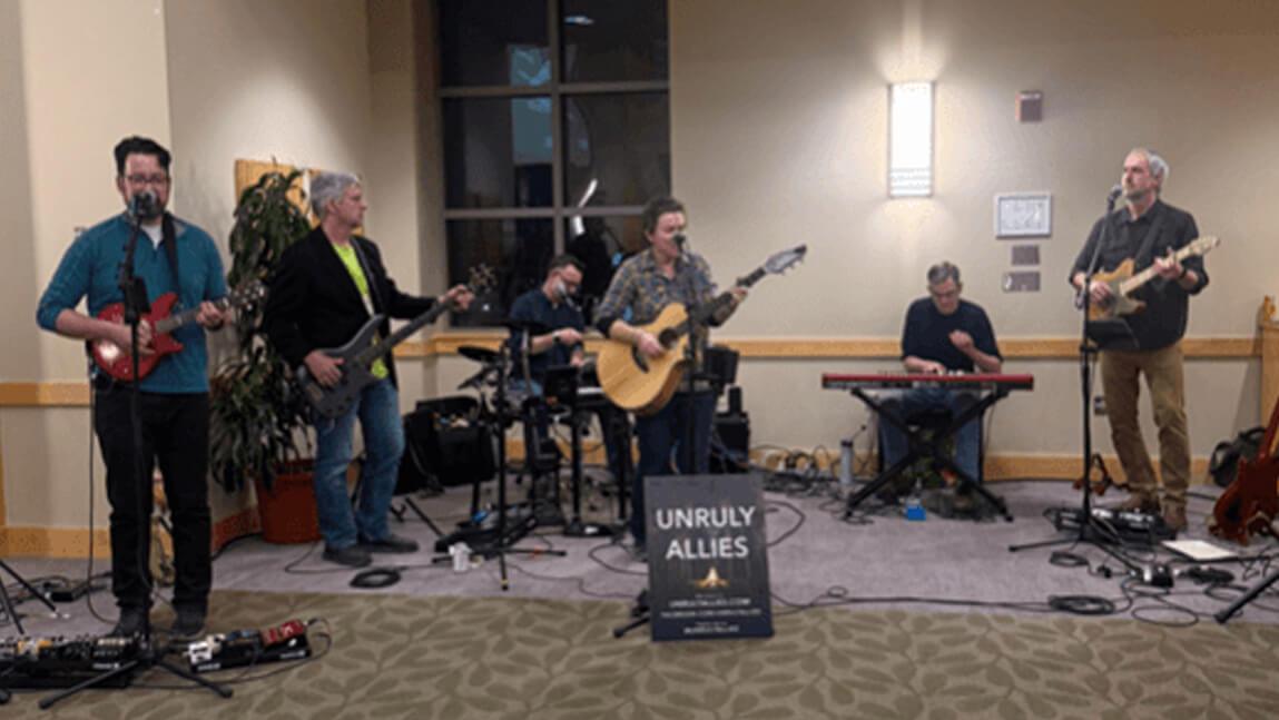 Six individuals in a band playing instruments behind a sign with the band name.