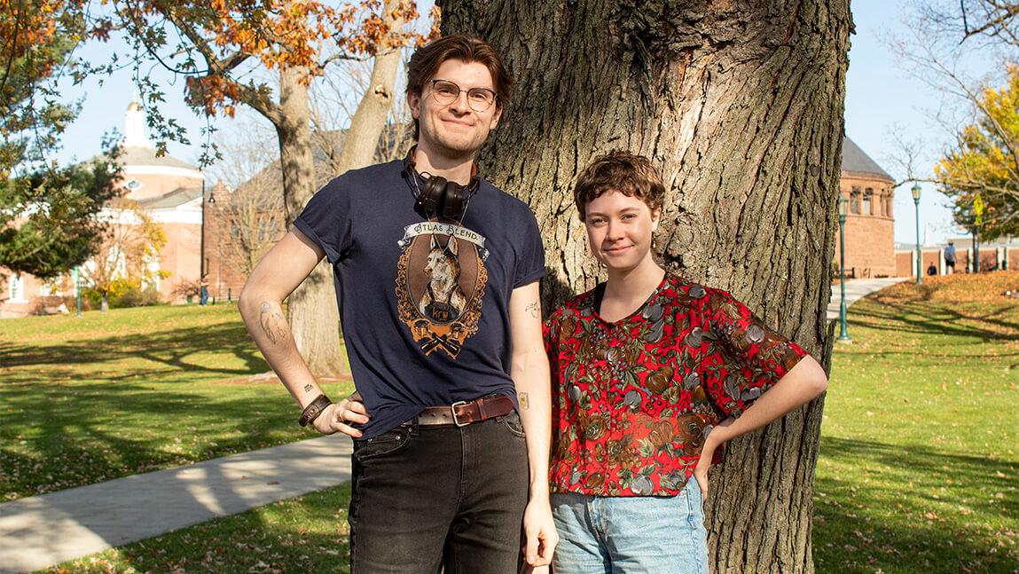 Two college students standing together in front of a large tree on the UVM campus with Billings Library in the background