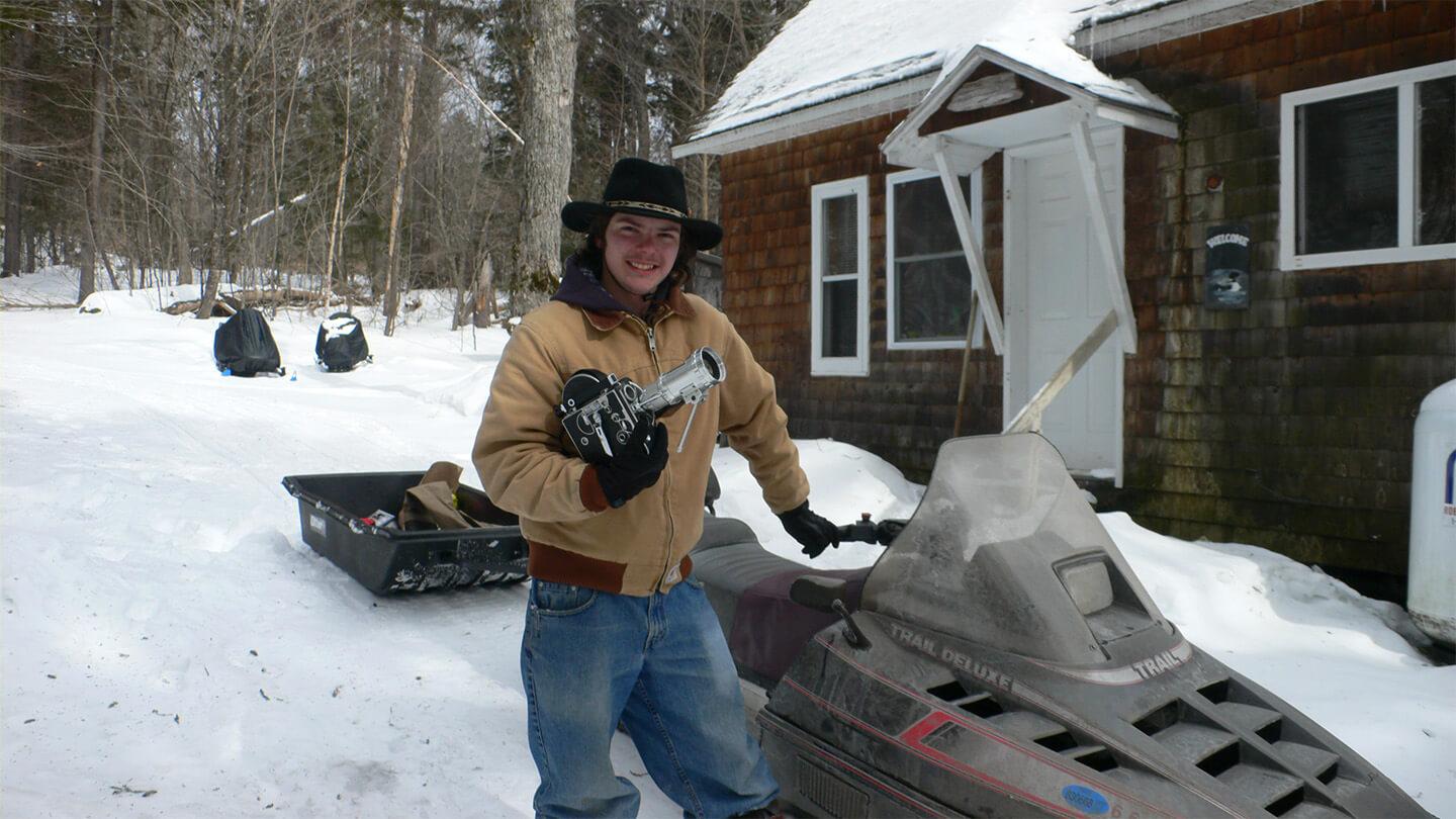young man with brimmed hat and tan jacket holding photo equipment and standing by snowmobile in front of snowy yard and red house