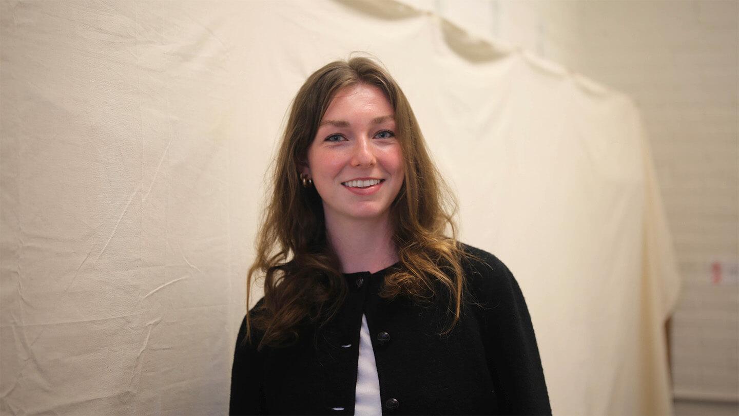 young woman with long light brown hair and black sweater over white shirt standing in front of canvas backdrop and smiling