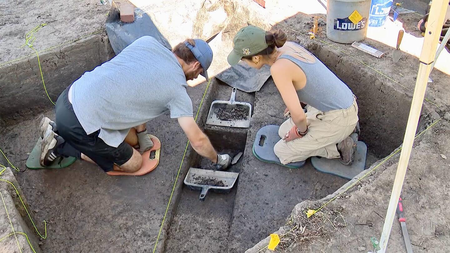 2 archaeologists (one male, one female) kneel in excavated area and work on soil analysis