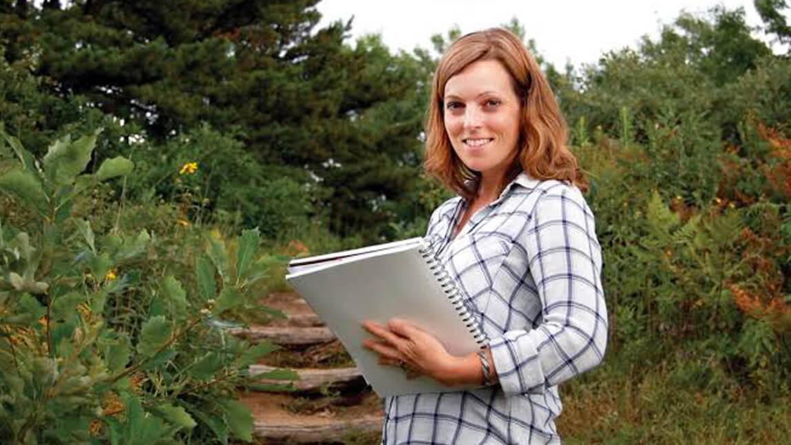 A person stands in the woods, holding a notebook