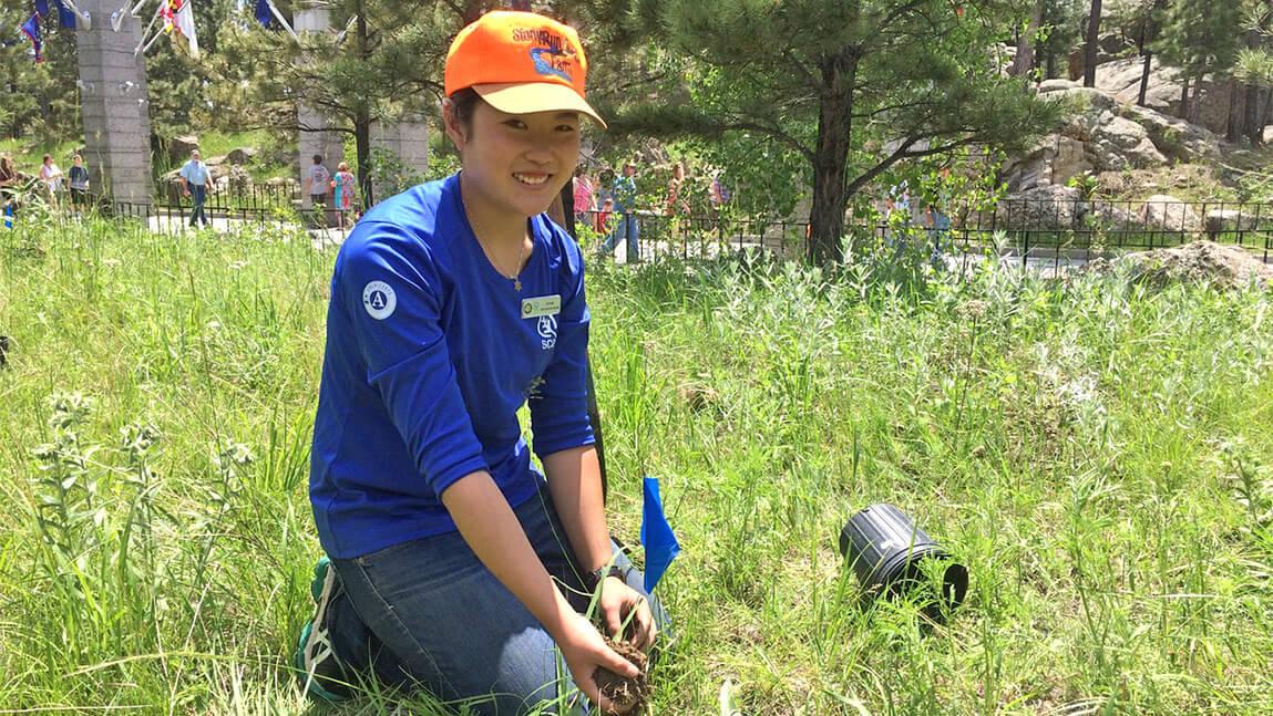 Amie Schiller in blue conservation shirt and orange hat kneels in grassy field to plant purple cone flower