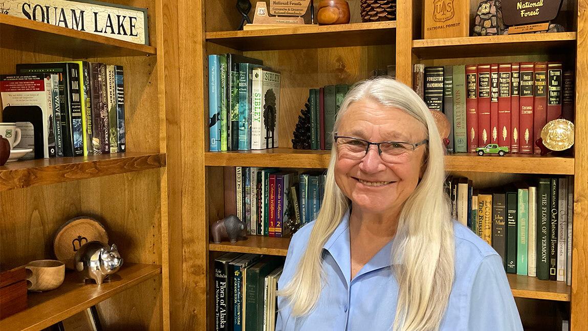 Gail Kimbell in front of wooden bookcases filled with Forestry books and memorabilia 