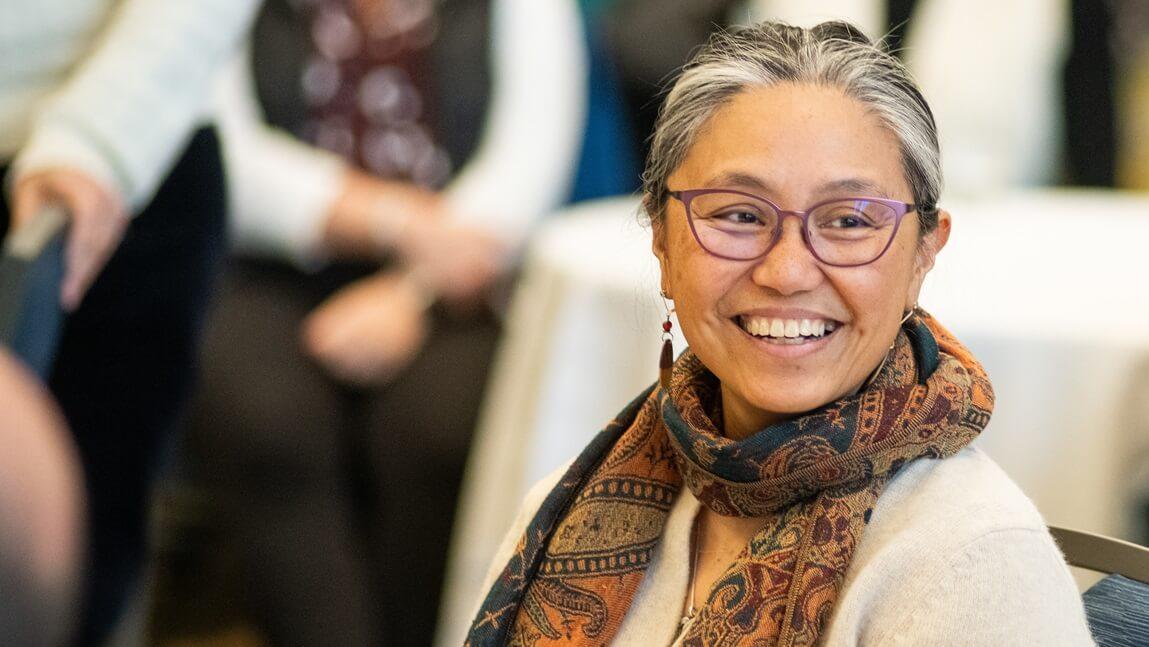 Marie Vea smiling during an event in the Davis Center. Vea is wearing glasses and a scarf.