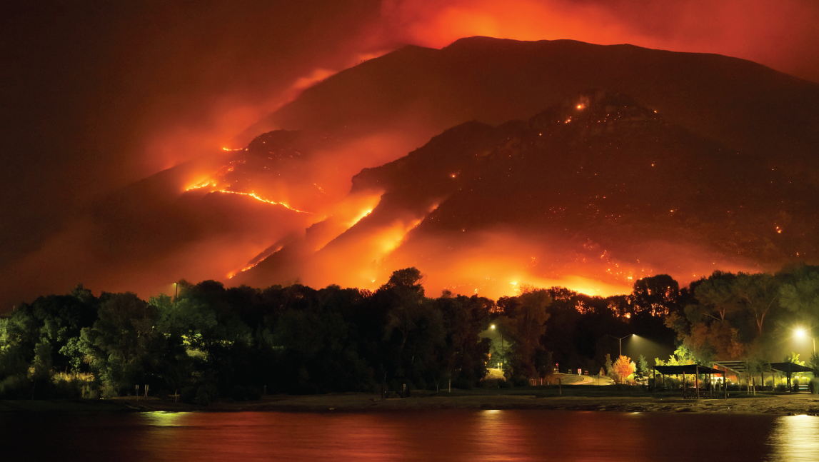 Image shows a forest fire in the background on mountains with a city and pond in the forefront. 