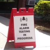 a bright red sign with words "Fire Alarm Testing in Progress" posted at building entrance